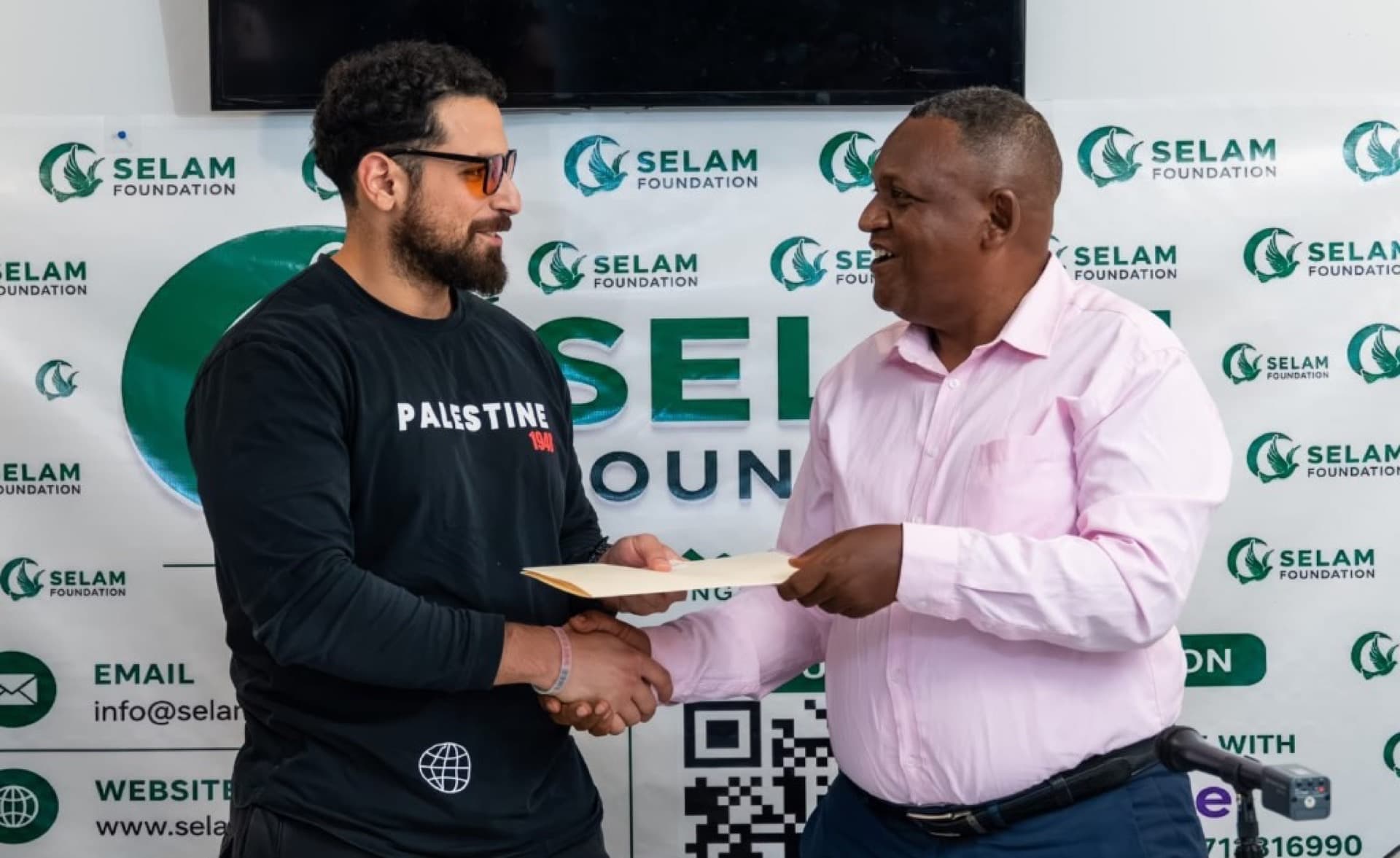 Two community members shake hands and exchange documents in front of a Selam Foundation banner